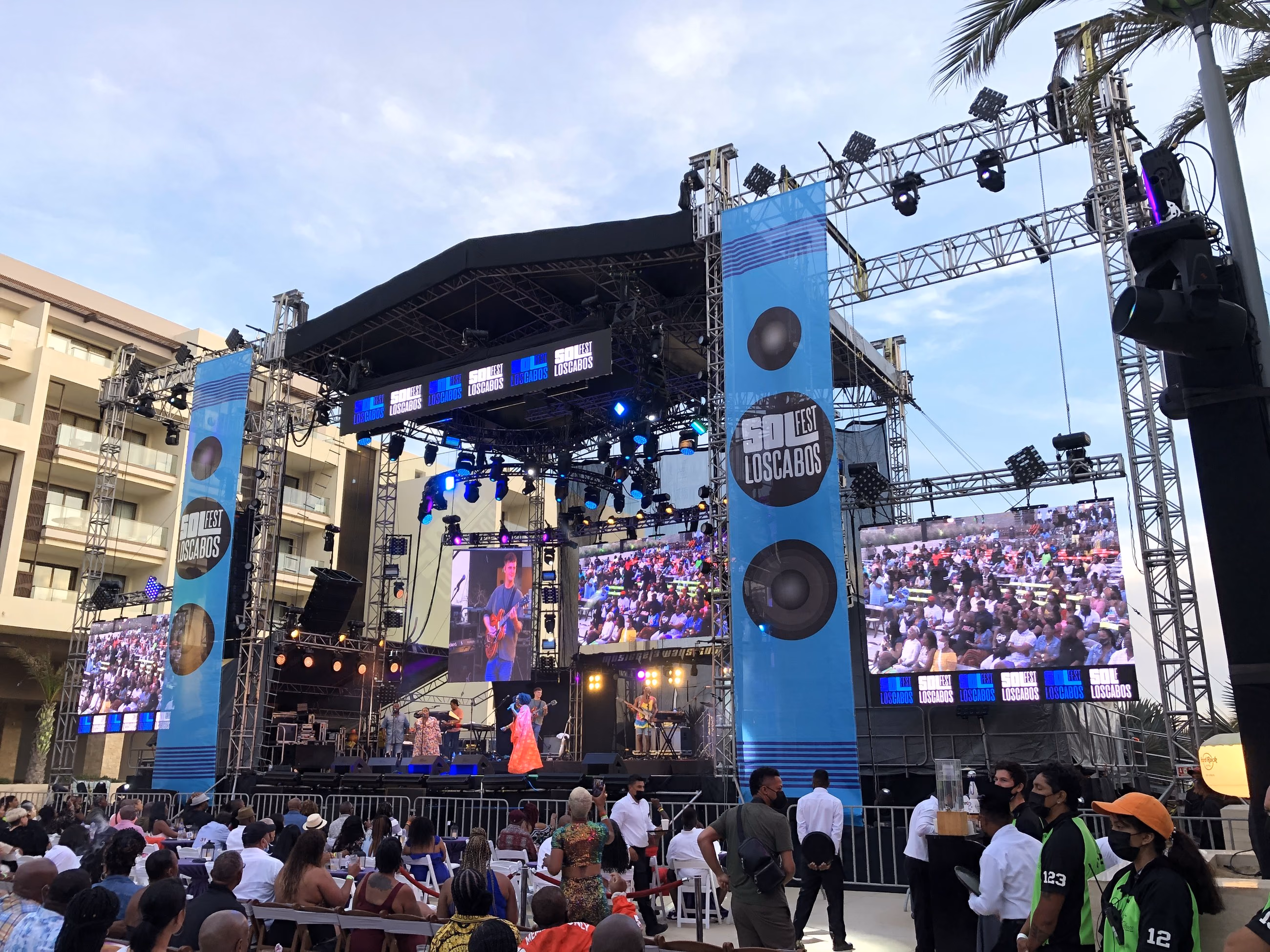 Escenario al aire libre con torres de audio, pantallas laterales y público de día