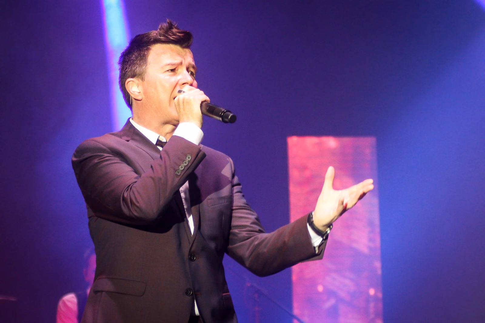 Rick Astley cantando en el escenario con pantalla de fondo en magenta